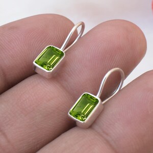 May include: A pair of silver drop earrings featuring rectangular, faceted lime green peridot gemstones set in smooth metal bezels with simple, curved wire hooks that extend upward from the top of each stone setting.