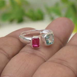 Puede incluir: Un anillo abierto de plata con dos piedras preciosas de corte rectangular. Una piedra es de color rosa vibrante y la otra es de color azul claro. La banda es de plata pulida y las piedras están engastadas en bisel, creando un diseño cruzado.