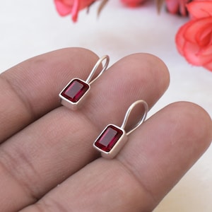 Può includere: Un paio di orecchini pendenti color argento con pietre rettangolari rosso scuro sfaccettate, incastonate in una montatura in metallo liscio. Ogni orecchino presenta un semplice gancio curvo, caratterizzato da un design pulito e rifinito.