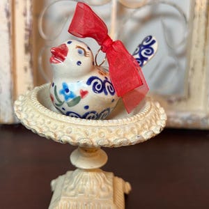 May include: A ceramic bird figurine with a red ribbon, perched on a decorative, off-white pedestal. The bird is painted with blue, red, and white floral patterns. The pedestal has a detailed, ornate design.