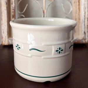 May include: A cream-colored ceramic container with a green band at the base and green floral and wave designs. The container has a slightly flared rim and a cylindrical shape. The background includes a decorative metal element.