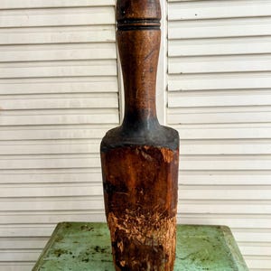 May include: A vintage wooden muddler with a dark brown, cylindrical head and a long, slender handle. The base is a rough-hewn block of wood, showing signs of age and use. The muddler is likely used for mixing cocktails or other beverages.