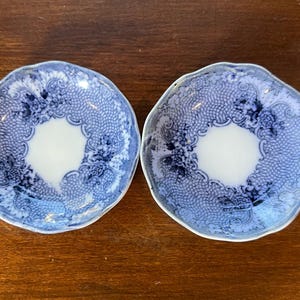May include: Two round ceramic saucers featuring a scalloped edge and a white center. The border is decorated with a blue and white intricate floral pattern and a fine mesh design that encircles the plain white basin of each small plate.