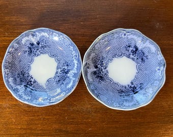 May include: Two round ceramic saucers featuring a scalloped edge and a white center. The border is decorated with a blue and white intricate floral pattern and a fine mesh design that encircles the plain white basin of each small plate.