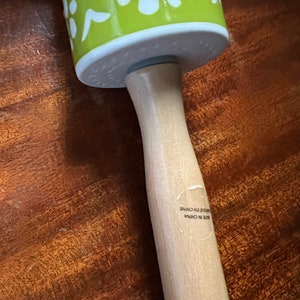 May include: A rolling pin with a light green barrel decorated with white floral patterns. The barrel is attached to a light-colored wooden handle. The text "MADE IN CHINA" is visible on the handle.