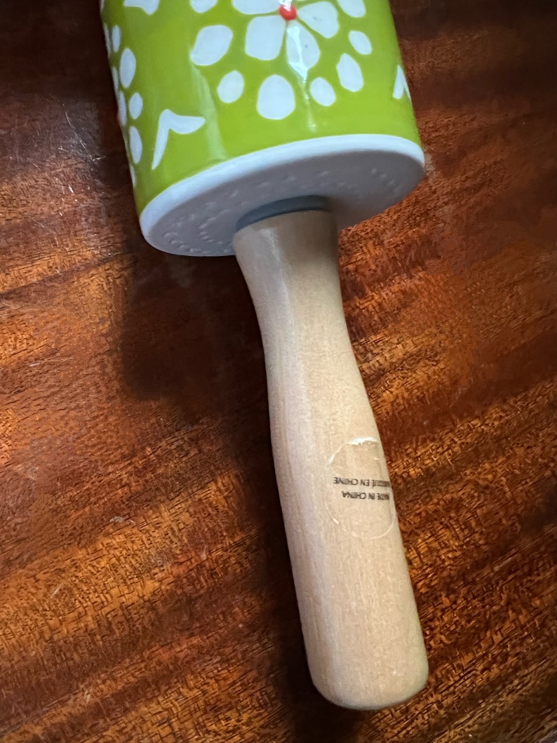 May include: A rolling pin with a light green barrel decorated with white floral patterns. The barrel is attached to a light-colored wooden handle. The text "MADE IN CHINA" is visible on the handle.