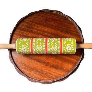 May include: A rolling pin with a green and white floral pattern and red stripes, with wooden handles. The rolling pin is resting on a round, wooden serving tray with a dark brown finish. Kitchenware.