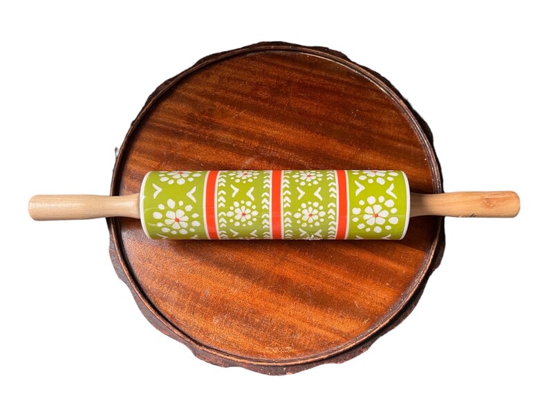 May include: A rolling pin with a green and white floral pattern and red stripes, with wooden handles. The rolling pin is resting on a round, wooden serving tray with a dark brown finish. Kitchenware.