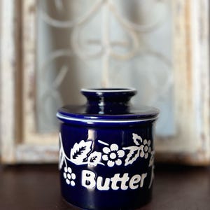 May include: A deep blue ceramic butter crock with a matching lid. The exterior features a white embossed floral pattern and the word Butter printed in a bold white font. The cylindrical container has a smooth, glossy finish and a white rim.