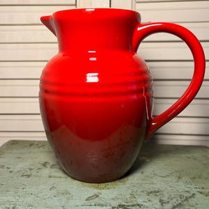 May include: A glossy red ceramic pitcher with a curved handle and a spout. The pitcher has horizontal ridges around the middle and a darker base. The pitcher is sitting on a distressed, light green surface.