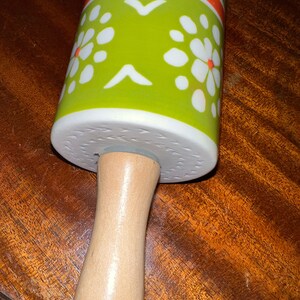 May include: A rolling pin with a wooden handle and a ceramic rolling surface. The ceramic surface is decorated with a green background, white floral patterns, and orange stripes. The rolling pin is designed for kitchen use.