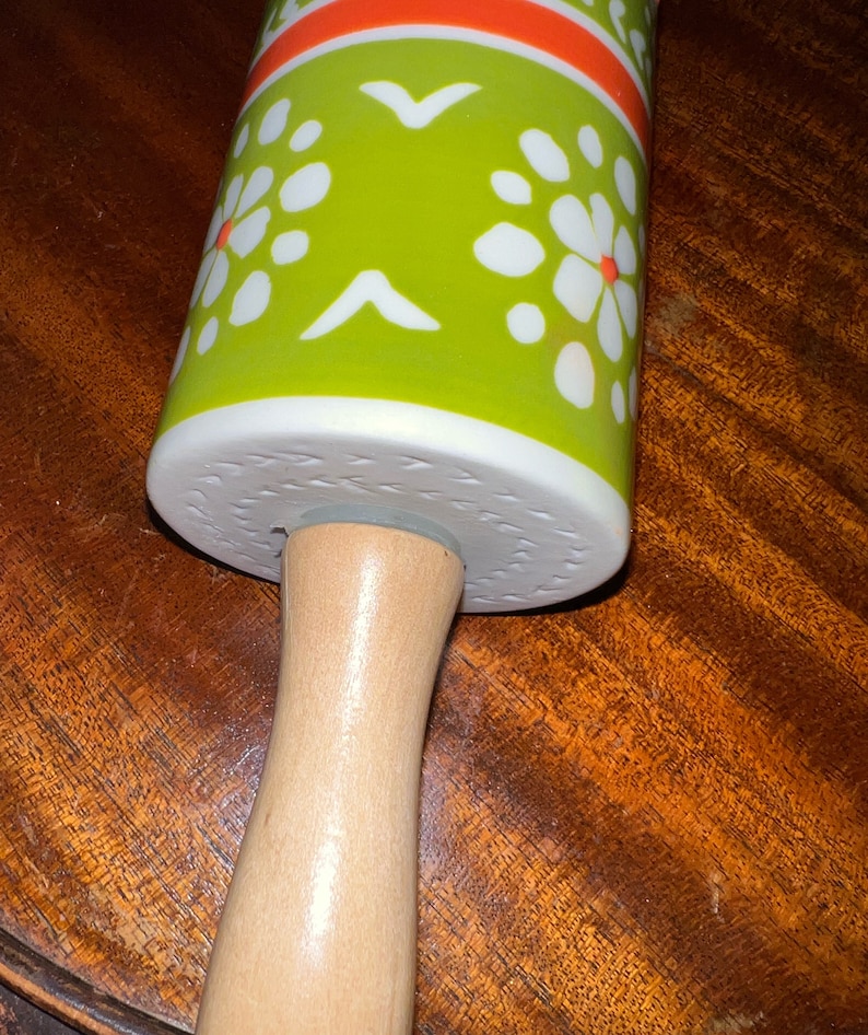 May include: A rolling pin with a wooden handle and a ceramic rolling surface. The ceramic surface is decorated with a green background, white floral patterns, and orange stripes. The rolling pin is designed for kitchen use.