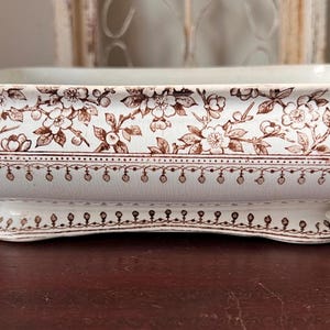 May include: A rectangular ceramic serving dish with a white base and brown transferware floral patterns. The exterior features a detailed botanical design of blossoms and branches, complemented by a decorative beaded border along the lower edge.