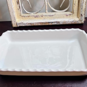 May include: A rectangular, off-white ceramic baking dish with scalloped edges. The dish has gently curved sides and is set against a dark background, with a decorative window frame in the background.