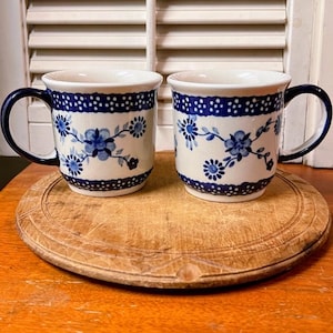 May include: Two ceramic mugs with blue floral patterns and dark blue handles. The mugs have a white base with blue floral designs and a dotted blue border. They sit on a round wooden cutting board.