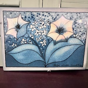 May include: Rectangular ceramic tile with a floral design. Two large, light-colored flowers with dark centers and blue leaves are featured. Small white flowers and a blue speckled background complete the design. The tile is framed in a silver metal.