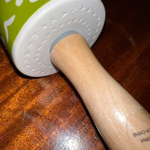 May include: A rolling pin with a wooden handle and a green and white patterned rolling surface. The rolling surface has a floral design with white flowers and green leaves. The handle is made of light-colored wood.