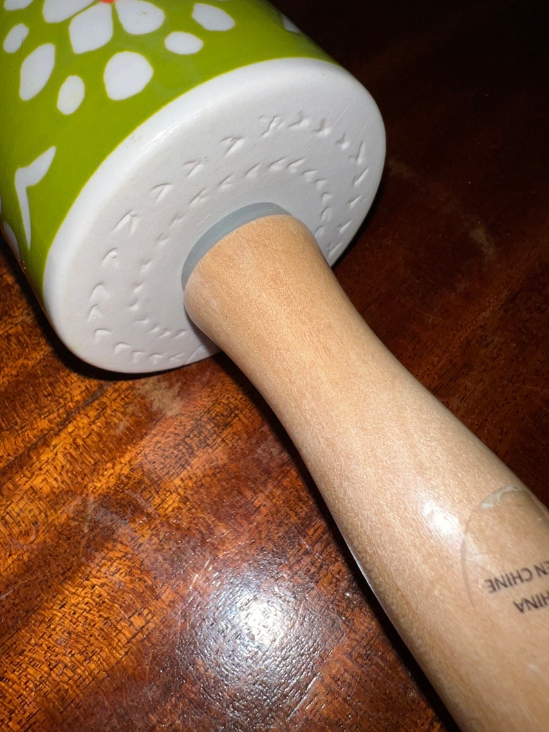 May include: A rolling pin with a wooden handle and a green and white patterned rolling surface. The rolling surface has a floral design with white flowers and green leaves. The handle is made of light-colored wood.