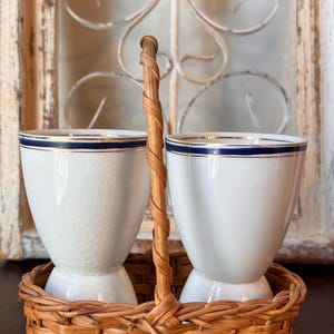 May include: Two white ceramic egg cups with a blue stripe and gold trim, presented in a woven wicker basket with a handle. The egg cups have a classic design, perfect for serving breakfast. The basket is a natural brown color.