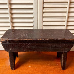 May include: A small, rectangular, dark brown wooden footstool with four short legs. The wood shows signs of wear and age, with visible grain and some chipped areas. The stool is set against a backdrop of white shutters.