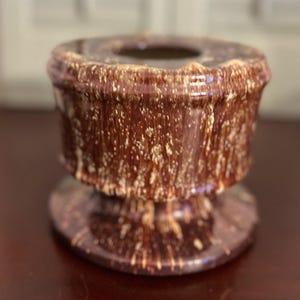 May include: A brown ceramic ashtray with a speckled, textured surface. The ashtray has a circular base and a cylindrical body with a flared top. The speckled pattern includes white and tan spots.
