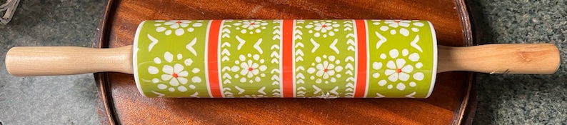 May include: A rolling pin with wooden handles and a patterned center. The center features a green background with white floral and geometric designs, accented by red vertical stripes. The handles are made of light-colored wood.