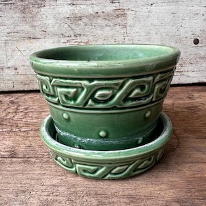 May include: A sage green ceramic plant pot with a matching saucer. The pot features a raised Greek key pattern around the upper rim and a row of small raised dots around the center. The glaze has a glossy finish with visible wear on the rim.