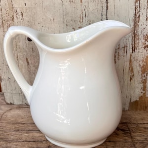 May include: A white ceramic pitcher with a smooth, glossy finish, featuring a curved handle on the left side and a wide, rounded pouring spout at the top. The vessel has a bulbous body that tapers toward the base, presenting a clean, solid white surface.