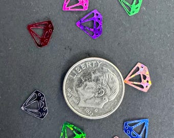 May include: A collection of small, colorful diamond-shaped embellishments in various colors, including pink, green, and blue. A U.S. dime is included for size comparison. The embellishments have a geometric design and are placed on a dark surface.