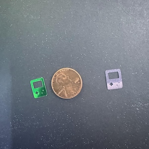 Puede incluir: Dos pequeños amuletos de consola de juegos brillantes, uno verde y otro morado, se muestran junto a un centavo. Los amuletos están detallados con una pantalla y botones, que recuerdan a un dispositivo de juego portátil clásico. El centavo muestra el perfil de Abraham Lincoln.