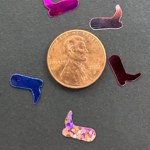 May include: A collection of colorful, miniature cowboy boot-shaped confetti scattered around a U.S. penny. The confetti pieces are in shades of pink, blue, and iridescent glitter. The penny is dated 2022.