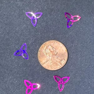May include: A collection of small, iridescent, three-leaf clover-shaped sequins in shades of purple and pink. A US penny is included for size reference. The sequins are scattered on a dark gray surface.