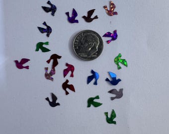 May include: A collection of iridescent bird-shaped sequins in various colors, including purple, green, and red, scattered around a U.S. dime. The sequins are small and reflective, creating a sparkling effect. The dime is silver and shows the profile of a person.
