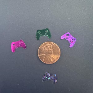 以下が含まれることがあります： ピンク、緑、青の色合いの、キラキラ光るゲームコントローラー型の装飾品のコレクション。ペニーを中心に配置されています。コントローラーは光沢のある反射面を持ち、濃いグレーの背景に散らばっています。