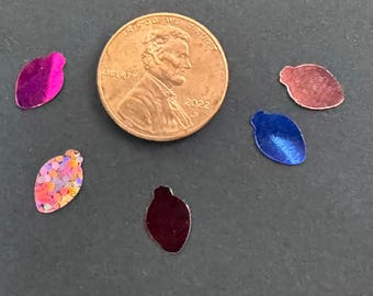 May include: A penny next to several small, leaf-shaped sequins in various colors. The sequins include pink, blue, red, and iridescent glitter. The penny is dated 2022.
