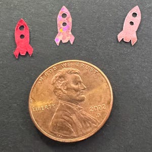 Può includere: Tre piccoli ritagli di razzi colorati, nelle tonalità del rosso e del rosa, sono esposti vicino a un penny. Il penny è datato 2022 e presenta il profilo di Abraham Lincoln. I razzi hanno finestre circolari e alette.