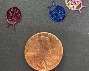 May include: Three ladybug-shaped embellishments in red, blue, and iridescent glitter, next to a copper penny. The ladybugs have cut-out details and are small in size. The penny shows the profile of Abraham Lincoln and the year 2022.