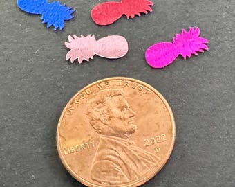 May include: Four small, colorful pineapple-shaped felt cutouts in blue, red, pink, and magenta, next to a 2022 penny. The felt pineapples have a textured appearance and are arranged on a dark gray surface.