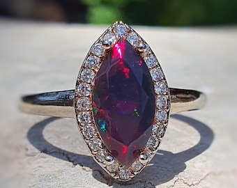 Op de afbeelding: Een ring met een marquise-geslepen edelsteen met een donkere, iriserende middensteen. De steen is omgeven door een halo van kleine, heldere diamanten. De band van de ring is dun en gepolijst goudkleurig.