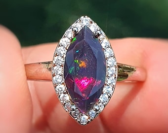 Op de afbeelding: Een gouden ring met een marquise-geslepen zwarte opaal edelsteen, omringd door een halo van kleine, heldere diamanten. De opaal vertoont flitsen van rood, groen en geel. De ring is geplaatst tegen een vage groene achtergrond.