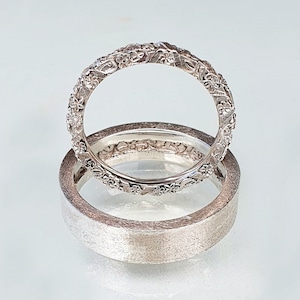 Puede incluir: Dos anillos de plata. El anillo superior presenta un diseño floral detallado, mientras que el anillo inferior tiene un acabado mate cepillado. Los anillos se muestran sobre una superficie reflectante, resaltando sus texturas y detalles intrincados. Estos anillos son probablemente para una boda o compromiso.