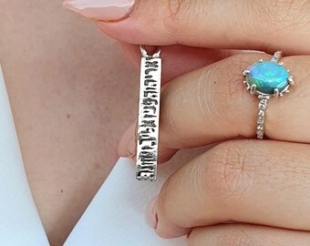 May include: A silver pendant necklace with a rectangular bar featuring engraved text. The pendant is held by a person. A ring with a blue opal stone is on the person's finger. The necklace is on a white shirt.