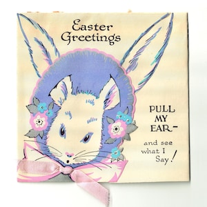 May include: A vintage Easter card featuring a white bunny with pink and blue flowers around its head. The bunny is wearing a purple hat and a pink bow. The text on the card reads "Easter Greetings" and "PULL MY EAR - and see what I Say!"