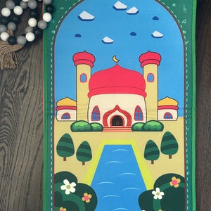 May include: A colorful prayer mat featuring a mosque design with a red dome and yellow minarets against a blue sky. The mat has a green border with musical notes and a pink trim. Decorative flowers and trees are also visible.