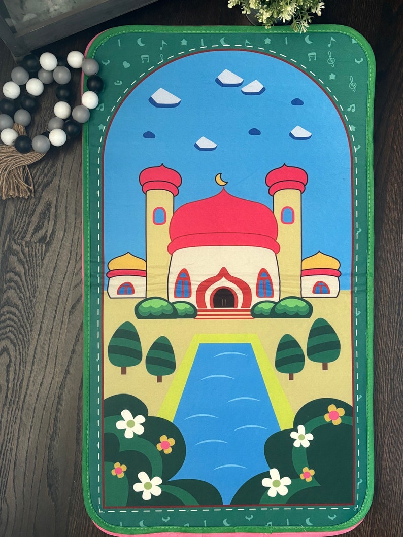 May include: A colorful prayer mat featuring a mosque design with a red dome and yellow minarets against a blue sky. The mat has a green border with musical notes and a pink trim. Decorative flowers and trees are also visible.