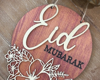 May include: A wooden round sign with the words "Eid Mubarak" in a white cursive font on a brown background. The sign is decorated with a white floral design.