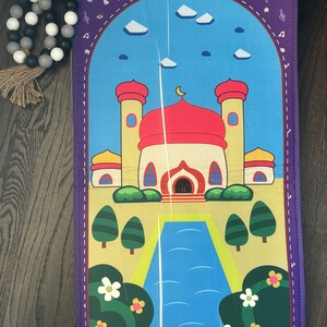 May include: A colorful prayer mat featuring a mosque design with a red dome, yellow walls, and blue sky. The mat has a purple border with musical notes and crescent moons. Decorative flowers and trees are also present.