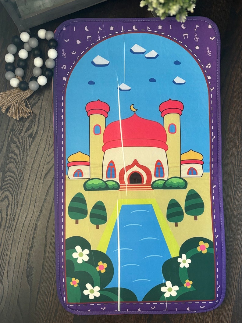 May include: A colorful prayer mat featuring a mosque design with a red dome, yellow walls, and blue sky. The mat has a purple border with musical notes and crescent moons. Decorative flowers and trees are also present.