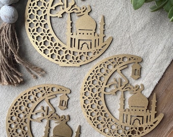 May include: Three gold-colored wooden cutouts of a crescent moon with a mosque and a lantern. The cutouts are on a beige fabric surface.