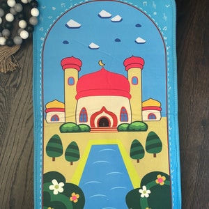 May include: A blue prayer mat featuring a cartoon mosque with red domes and yellow minarets. The design includes a blue sky with white clouds, a crescent moon, and musical notes. Green trees and flowers frame a blue river.